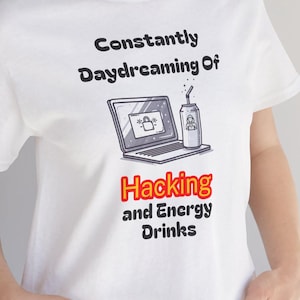 May include: White t-shirt with the text "Constantly Daydreaming Of Hacking and Energy Drinks." The graphic features a laptop and a can of energy drink. The shirt is a casual wear item.