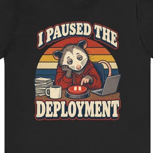 May include: A black t-shirt featuring a graphic of an opossum in a red hoodie sitting at a desk with a laptop, a mug, and a stack of papers. The opossum presses a red pause button. The text reads I PAUSED THE DEPLOYMENT in a retro font.