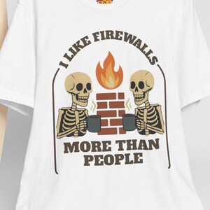 Puede incluir: Camiseta blanca con un gráfico humorístico. El diseño presenta dos figuras de esqueleto sosteniendo tazas, una chimenea de ladrillo y una llama, con el texto "I like firewalls more than people."