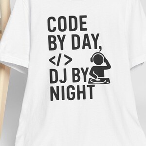 Peut inclure: T-shirt blanc avec l'inscription noire « CODE BY DAY, </> DJ BY NIGHT ». Le motif comprend un graphisme de DJ. Une casquette de baseball blanche repose sur une chaise en bois derrière le t-shirt.