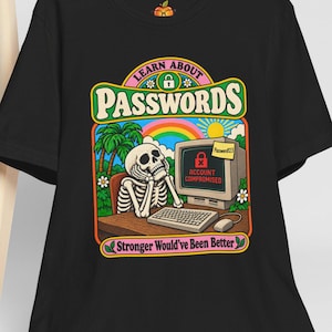 May include: A black t-shirt featuring a colorful graphic of a skeleton sitting at a computer with the text "LEARN ABOUT PASSWORDS" and "Stronger Would've Been Better". The screen displays "ACCOUNT COMPROMISED" with a lock icon and a sticky note.