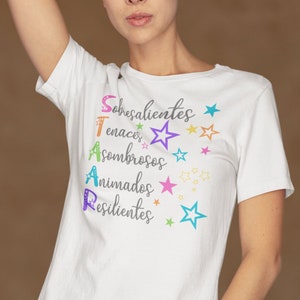 Puede incluir: Camiseta blanca con un diseño gráfico colorido que deletrea "STAR" con las palabras "Sobresalientes", "Tenaces", "Asombrosos", "Animados" y "Resilientes" escritas verticalmente junto a cada letra.