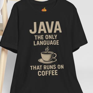 May include: Black t-shirt with the text "JAVA THE ONLY LANGUAGE THAT RUNS ON COFFEE" in beige. The design includes a coffee cup graphic. A white baseball cap rests on a wooden chair in the background.
