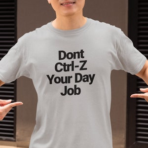Op de afbeelding: Een lichtgrijs T-shirt met zwarte tekst die "Dont Ctrl-Z Your Day Job" zegt.