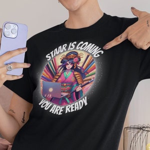 May include: A black t-shirt featuring a colourful graphic of a woman in a kimono holding a laptop. The graphic includes the text "Staar is coming you are ready".
