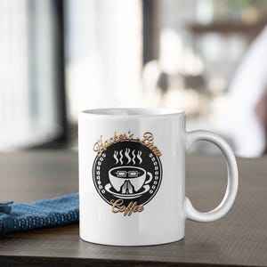 Pode incluir: Caneca de café de cerâmica branca com um design preto e dourado. O design apresenta as palavras "Hacker's Brew Coffee" em torno de um gráfico de xícara de café com vapor subindo. A caneca tem uma alça curva e está em uma superfície de madeira.