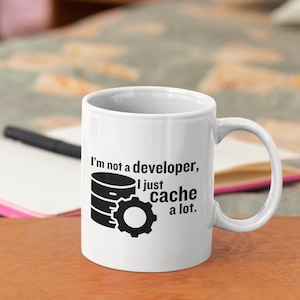 Op de afbeelding: Witte keramische mok met een zwart handvat. De mok heeft de tekst "I'm not a developer, I just cache a lot." samen met een afbeelding van een database en een tandwiel. De mok staat op een houten oppervlak.