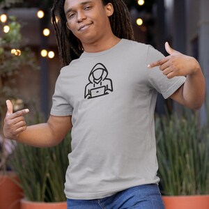 May include: A gray t-shirt with a black graphic of a person wearing a hoodie and using a laptop. The person's face is not visible.