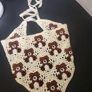May include: Cream-colored crochet halter top featuring a repeating pattern of brown teddy bears. The top has a triangular shape with a tie closure at the neck. The open weave design adds a delicate touch to this handmade garment.