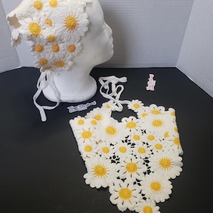 May include: A white crochet hat and neck scarf set, adorned with yellow-centered daisies. The hat is on a mannequin head, and the scarf is laid out below. The set is handmade and features a delicate, floral design.