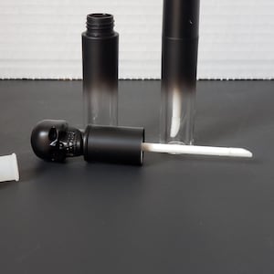 May include: Two black lip gloss tubes with a skull-shaped top. One tube is open and the applicator wand is lying on a black surface.