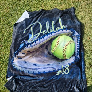 May include: A black fleece blanket with a personalized design featuring a softball in a catcher's mitt. The name "Delilah" and the number "#18" are printed on the blanket.