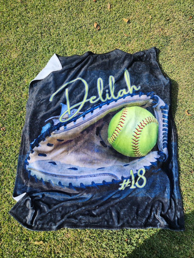 May include: A black fleece blanket with a personalized design featuring a softball in a catcher's mitt. The name "Delilah" and the number "#18" are printed on the blanket.