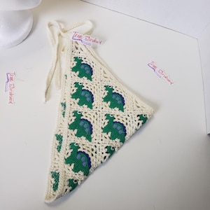 May include: Cream-colored, crocheted bandana featuring a repeating pattern of green and blue turtles. The bandana has a triangular shape with a drawstring closure. The text "True Storyhand" is printed on small labels.