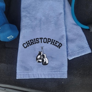 May include: A light blue towel with the name "CHRISTOPHER" in black, arched text. Below the name are two black and white boxing gloves. A blue dumbbell and exercise band are in the background.