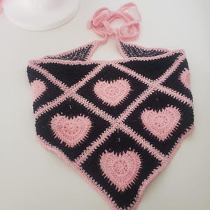 May include: A handmade crochet top in a triangular shape, featuring a black and pink heart pattern. The top has a halter neck tie and a pink border. The design includes multiple pink hearts on a black background.