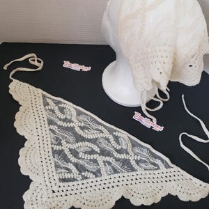 May include: Ivory-colored, crocheted baby bonnet and shawl set. The bonnet is displayed on a white mannequin head. The shawl is triangular with a scalloped edge and a delicate, openwork design. The set is made of a soft, lightweight material.