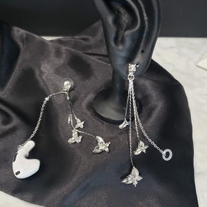 May include: A pair of silver dangle earrings with butterfly charms. The earrings are on a black fabric background.