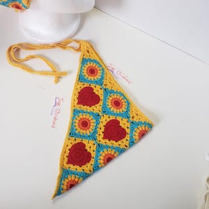 May include: A handmade crochet triangle top featuring a vibrant pattern of red hearts and sunflowers on a yellow and turquoise background. The top has a yellow tie for an adjustable fit. The overall design is a colorful and cheerful summer accessory.