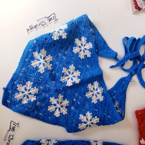 May include: A blue crocheted scarf featuring white snowflake designs. The scarf has a triangular shape with ties at the ends. The True Story logo is visible in the image.