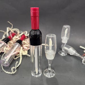 May include: Clear lip gloss tubes with red and black caps, and a clear champagne glass-shaped lip gloss applicator. The lip gloss tubes are arranged on a dark surface, with a few tubes and applicators scattered around.