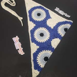 May include: A handmade crochet bandana in a triangular shape. The bandana features a white base with blue and light blue eye-shaped designs. The bandana has long white ties for securing around the neck. The text "True Storyboard" is visible on a sticker.