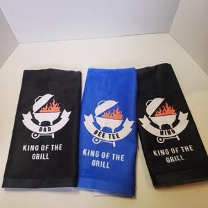 May include: Three kitchen towels with a grilling theme. One black, one blue, and one black towel feature a grill graphic with flames and the words "DAD", "DEE TEE", and "NINO" above "KING OF THE GRILL".