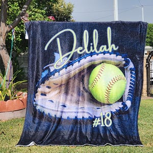 May include: A black fleece blanket with a personalized name "Delilah" and the number "#18" printed on it. The blanket features a graphic of a softball in a catcher's mitt.