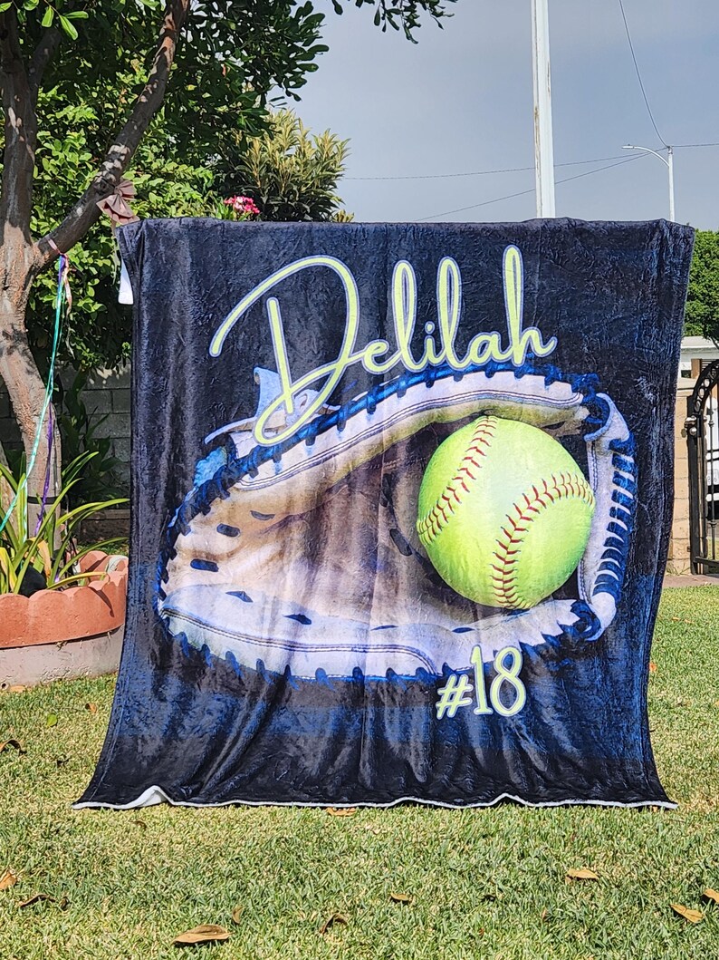 May include: A black fleece blanket with a personalized name "Delilah" and the number "#18" printed on it. The blanket features a graphic of a softball in a catcher's mitt.