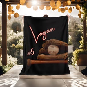 May include: Black fleece blanket with a red baseball glove, a baseball, and a wooden bat. The text "Logan #5" is printed in red on the blanket.