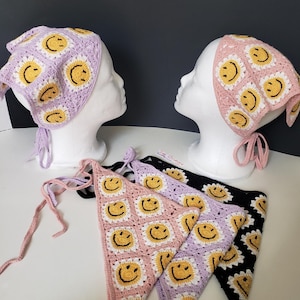 May include: Crocheted bandanas in various colors, including pink, lavender, and black, each adorned with a repeating smiley face pattern. The bandanas are displayed on mannequin heads and laid flat, showcasing their design and tie closures.
