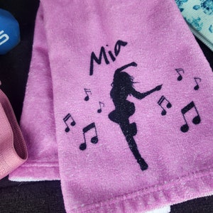 May include: Pink towel with a white border, featuring a black silhouette of a dancer with musical notes and the name "Mia" embroidered on it.