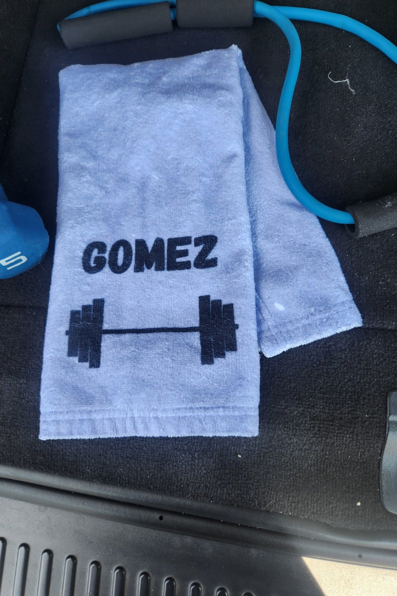 Puede incluir: Una toalla azul claro con el nombre "GOMEZ" y un dise&ntilde;o de barra de pesas negra.