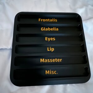 May include: Black tray with six compartments labeled "Frontalis", "Glabella", "Eyes", "Lip", "Masseter", and "Misc."