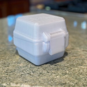 May include: A small, white, plastic box with a hinged lid and a latch. The box is made of a textured material and has a slightly rounded shape.