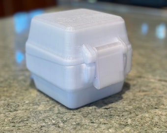 Puede incluir: Una pequeña caja de plástico blanca con una tapa con bisagras y un cierre. La caja está hecha de un material texturizado y tiene una forma ligeramente redondeada.