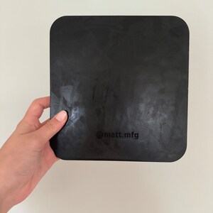 May include: A black square-shaped cutting board with rounded corners. The board has a textured surface and the text "@matt_mfg" is printed on the bottom.