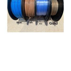 May include: Four spools of filament for 3D printing. The spools are labeled "Blue", "Tan", "translucent blue", and "Gold".