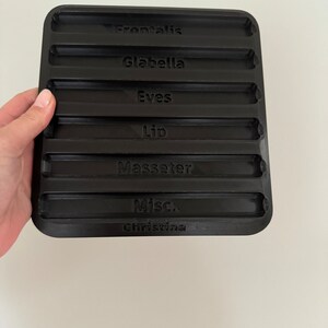 May include: Black plastic tray with eight indented slots labeled "Frontalis", "Glabella", "Eves", "Lip", "Masseter", "Misc.", and "Christina".