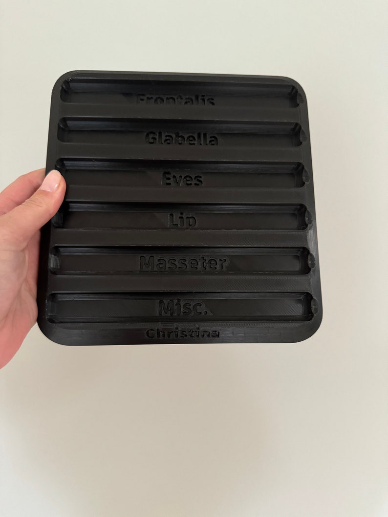 May include: Black plastic tray with eight indented slots labeled "Frontalis", "Glabella", "Eves", "Lip", "Masseter", "Misc.", and "Christina".