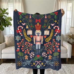 May include: A navy blue Christmas-themed blanket featuring a central nutcracker figure in a red and pink uniform. The design includes candy canes, snowflakes, gingerbread men, and gift boxes in red, white, and gold. The blanket is held up by a person.