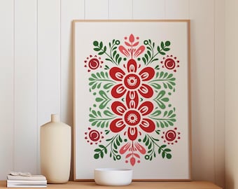 May include: A framed art print featuring a symmetrical floral design in red, green, and pink on a cream background. The artwork is displayed on a wooden table with a vase and a white bowl.