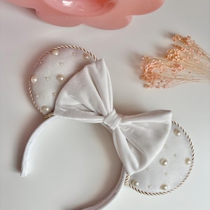May include: White velvet mouse ear headband with a large bow. The ears are decorated with faux pearls and a gold rope trim. A pink decorative object is in the background.
