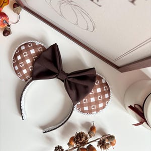 May include: A headband featuring two circular ears with a brown and white gingham pattern, accented by small Mickey Mouse head silhouettes. A large, solid dark brown fabric bow is centred between the ears, and the headband edges are trimmed with white rope.