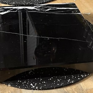 May include: A black marble square tabletop with white veining. The tabletop is on a black and white speckled surface.