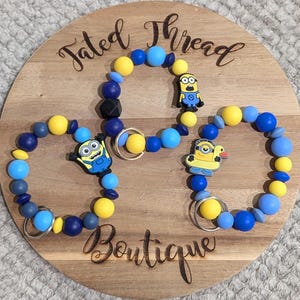 Puede incluir: Tres pulseras de cuentas azules y amarillas, cada una con un dije de personaje Minion. Las pulseras están dispuestas sobre una tabla de madera con el texto "Fated Thread Boutique".