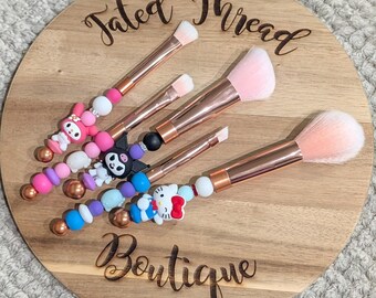 May include: A set of five makeup brushes with rose gold handles and pink bristles. The handles are decorated with colorful beads and character charms, including My Melody, Keroppi, and Hello Kitty. The brushes are arranged on a wooden board with the text "Tated Thread Boutique".
