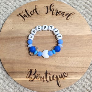May include: A blue and white beaded bracelet with the word "SWIFTIE" spelled out in white letter beads. The bracelet has a silver-colored key ring and is on a wooden round board with the words "Tated Thread Boutique" engraved on it.