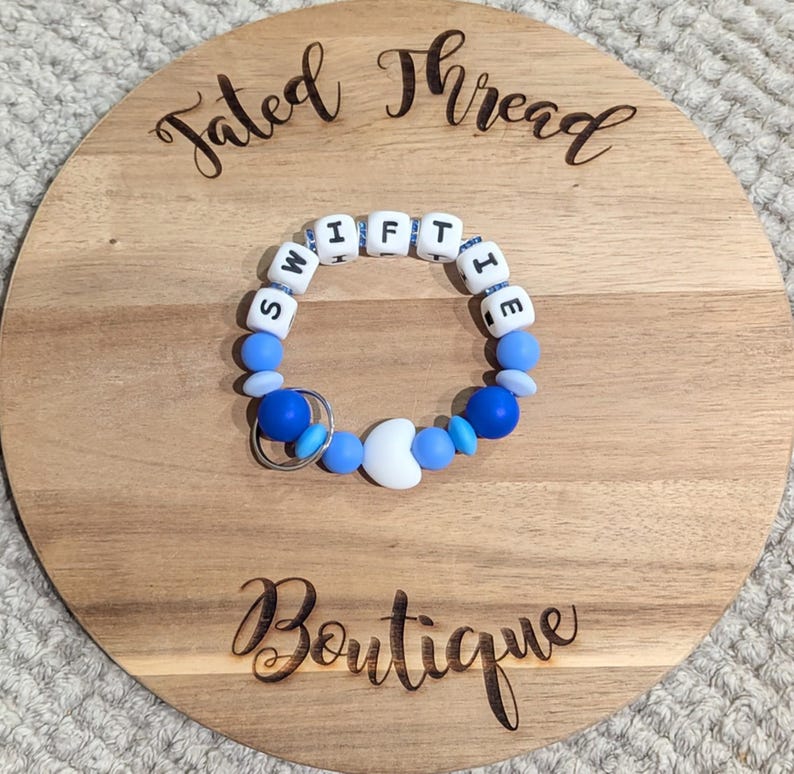 May include: A blue and white beaded bracelet with the word "SWIFTIE" spelled out in white letter beads. The bracelet has a silver-colored key ring and is on a wooden round board with the words "Tated Thread Boutique" engraved on it.