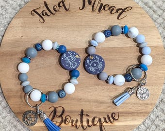 May include: Two beaded keychains with zodiac sign charms. The keychains feature white, gray, and blue beads, with a blue tassel and a silver charm. The zodiac signs are on dark blue circular pendants. The keychains are displayed on a wooden board with the text "Tated Thread Boutique".